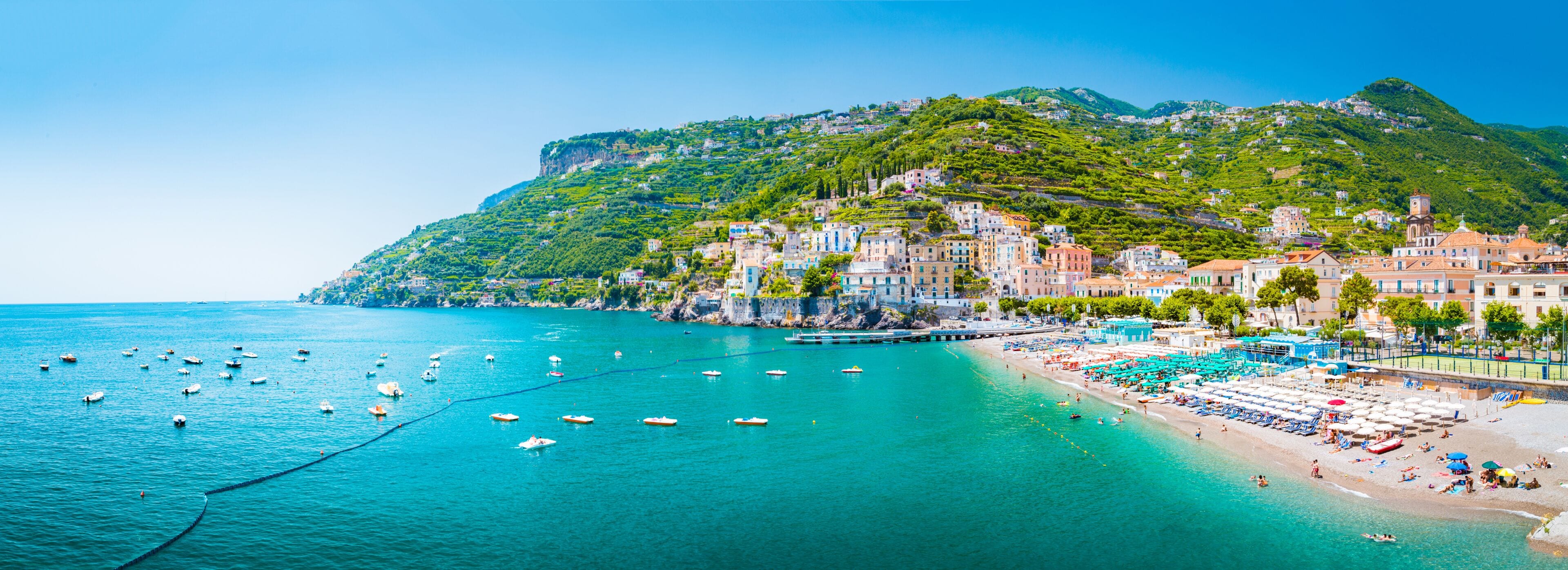 A coastal town with a hillside, boats on the water, and beach umbrellas.