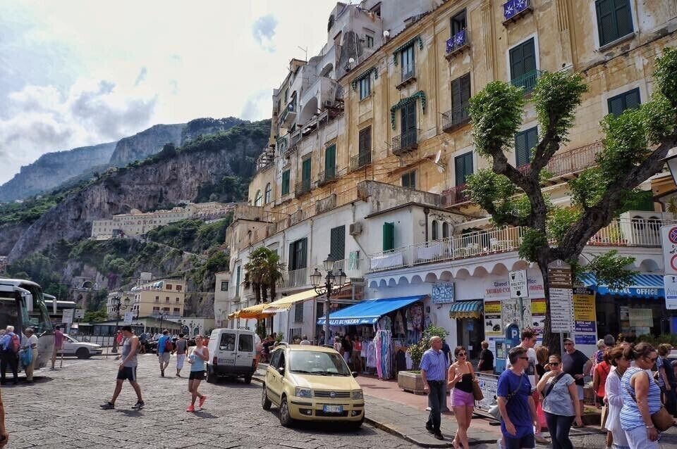 Uma rua de paralelepípedos com pessoas caminhando, carros estacionados e edifícios com sacadas e lojas.