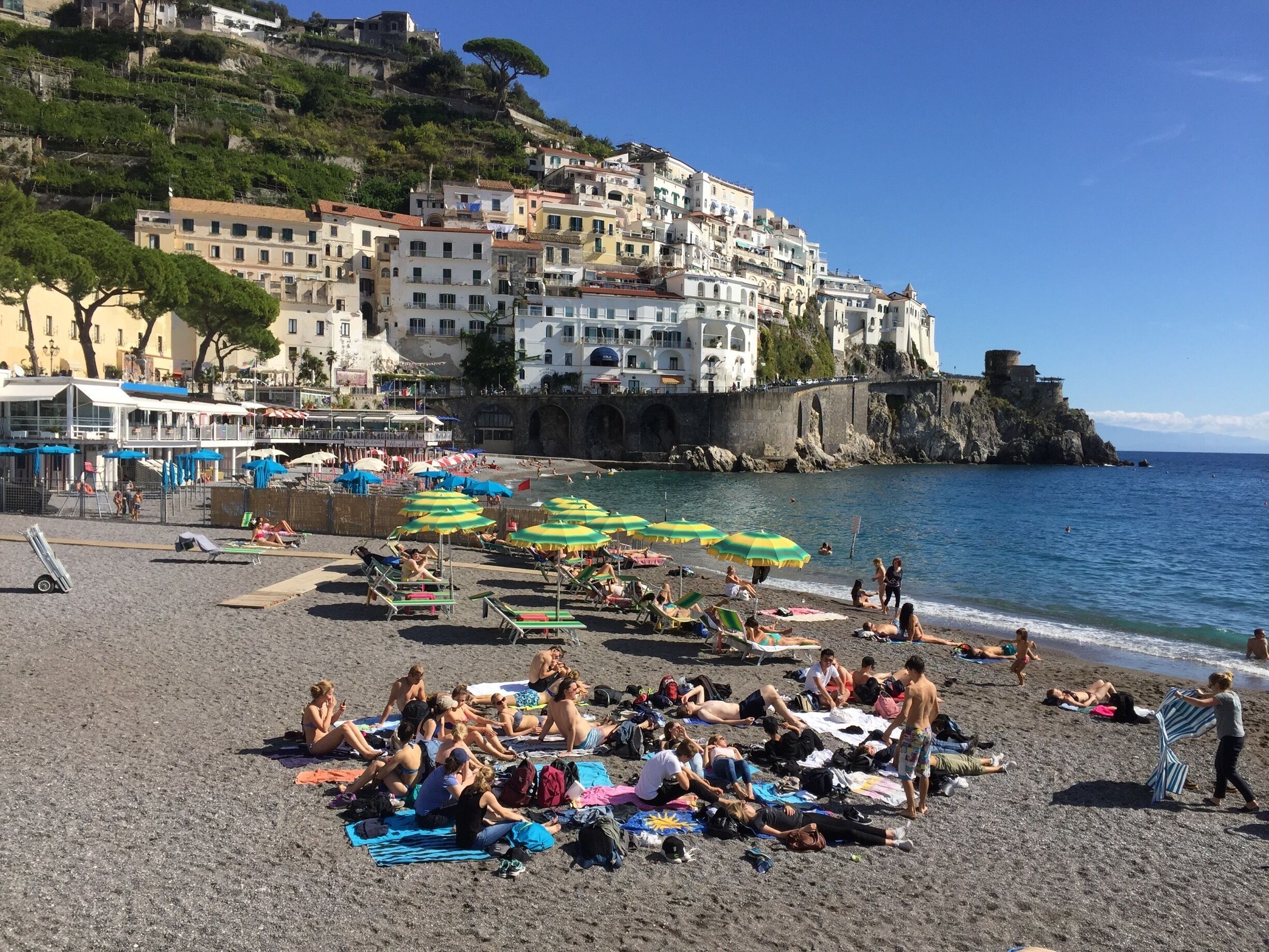 Uma praia movimentada com pessoas se banhando ao sol e nadando, cercada por edifícios coloridos e um castelo em um penhasco.