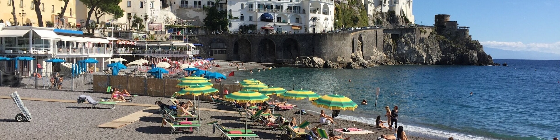 Uma praia movimentada com pessoas se banhando ao sol e nadando, cercada por edifícios coloridos e um castelo em um penhasco.