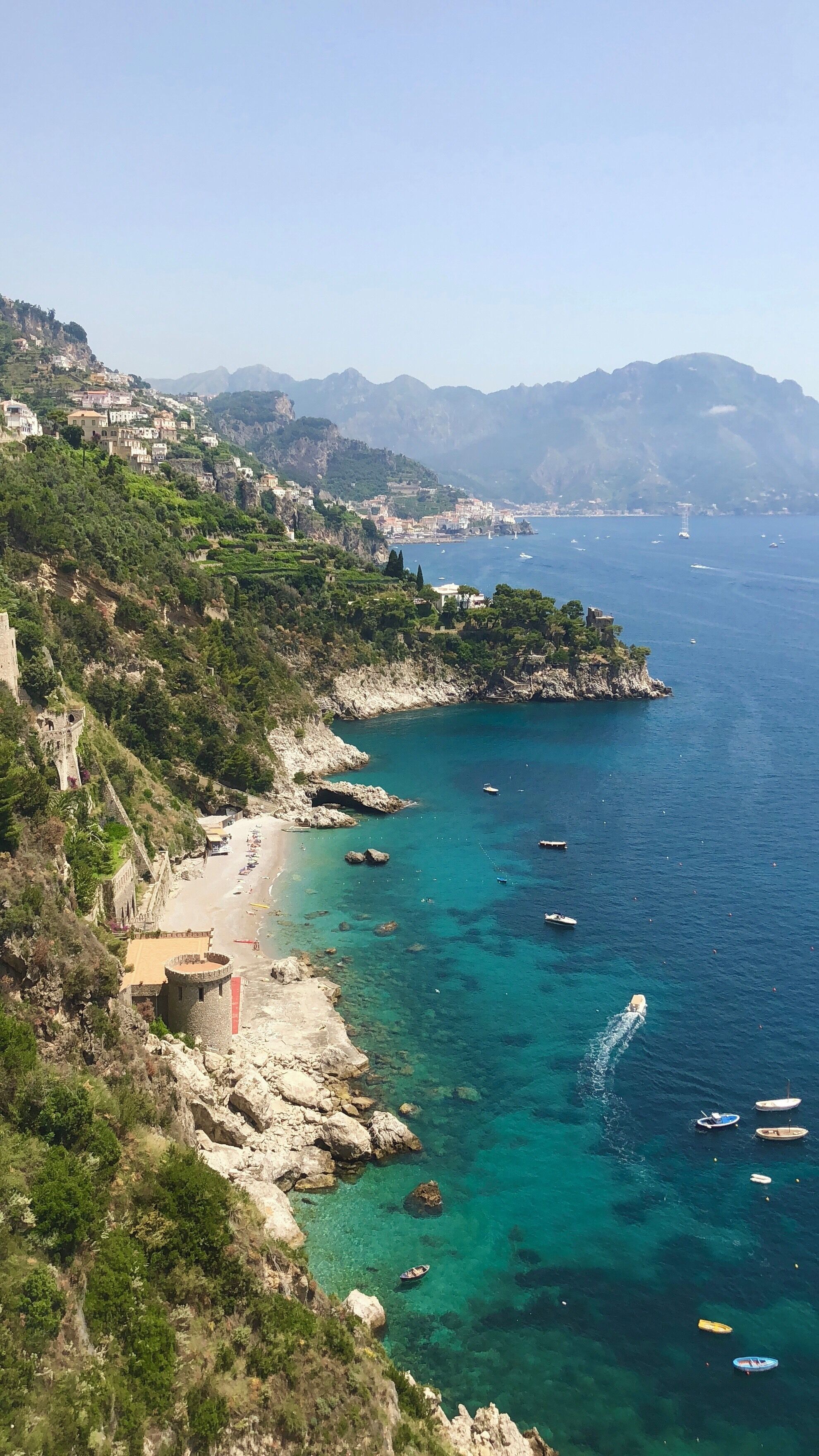 A coastal landscape with a clear blue sea, rocky cliffs, and a beach with boats.