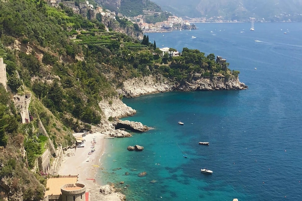 A coastal landscape with a clear blue sea, rocky cliffs, and a beach with boats.