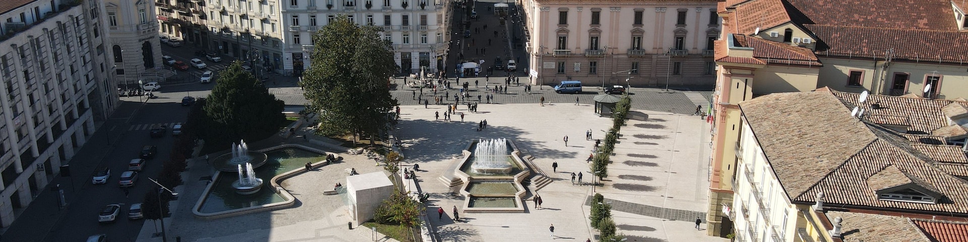 A square with fountains, surrounded by historic buildings and a mountainous backdrop.