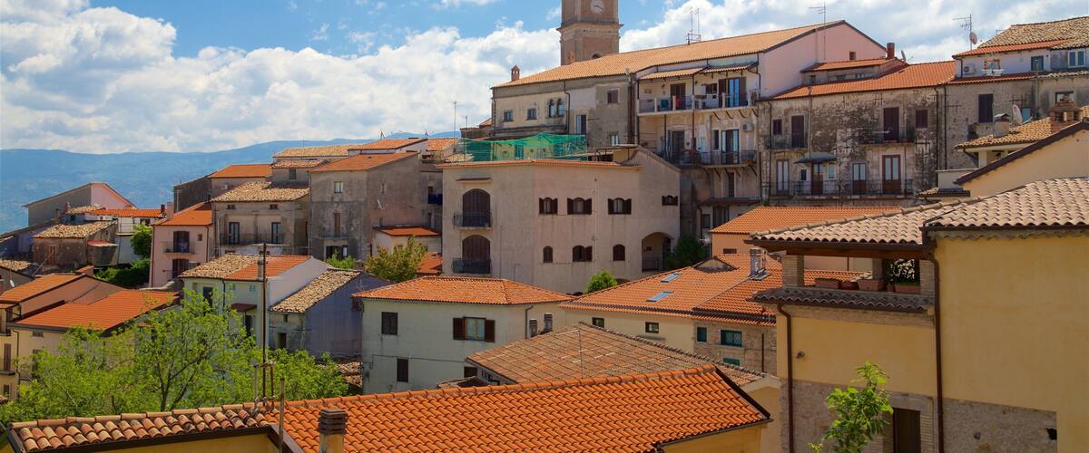A town with terracotta-roofed buildings, a prominent clock tower, and a mountainous backdrop.