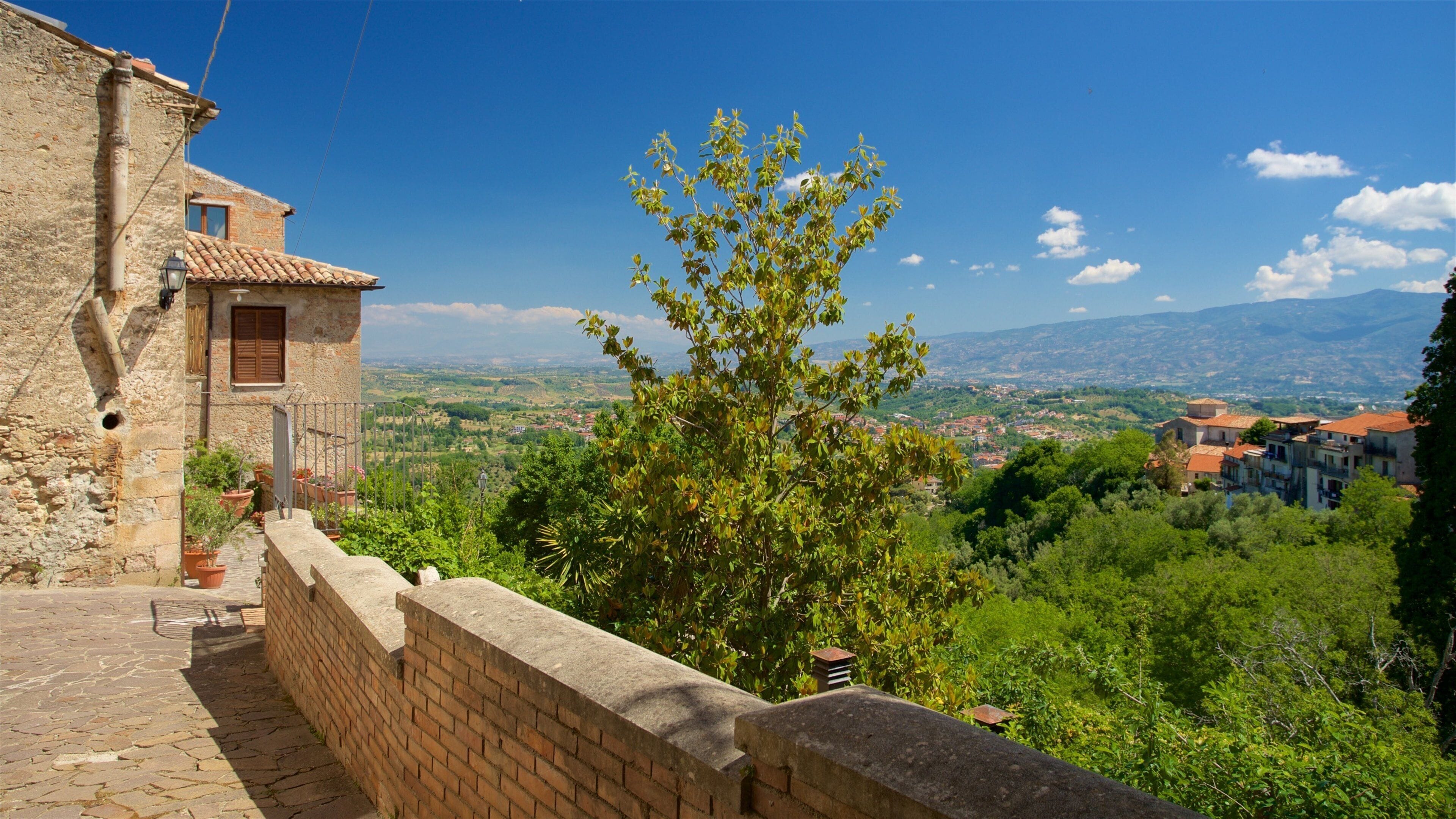 A stone building with a tiled roof overlooking a valley with greenery and distant mountains.