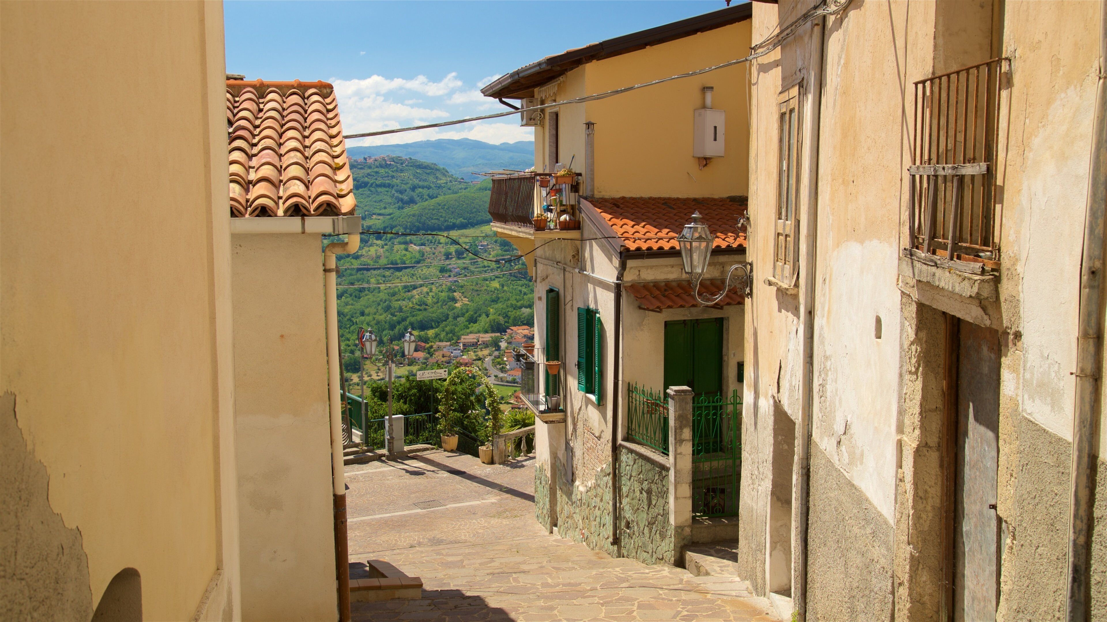 A narrow street with terracotta-tiled roofs, green shutters, and a stone staircase leading to a scenic hillside view.