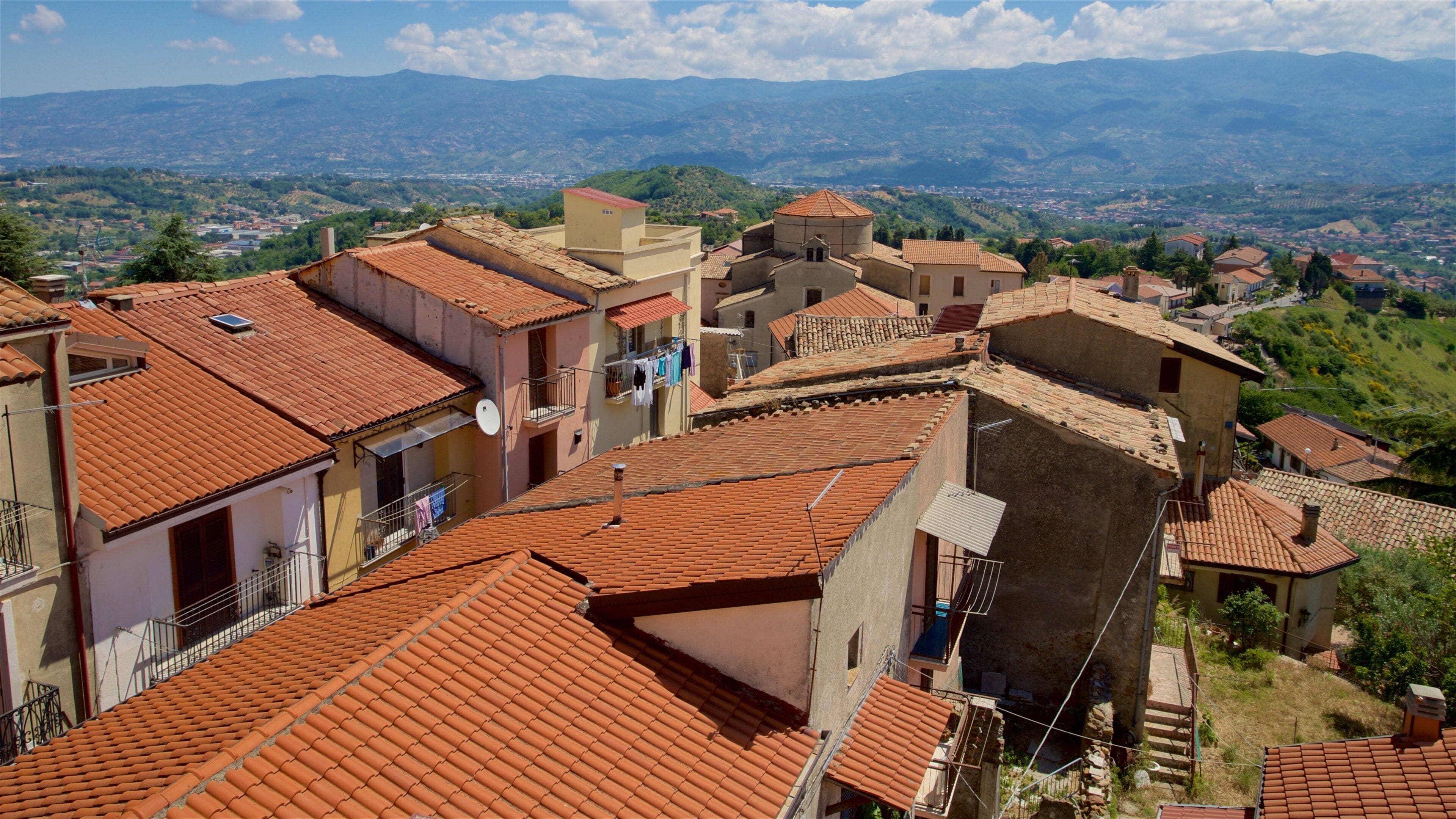 A town with terracotta-roofed buildings, a mountainous backdrop, and a clear sky.