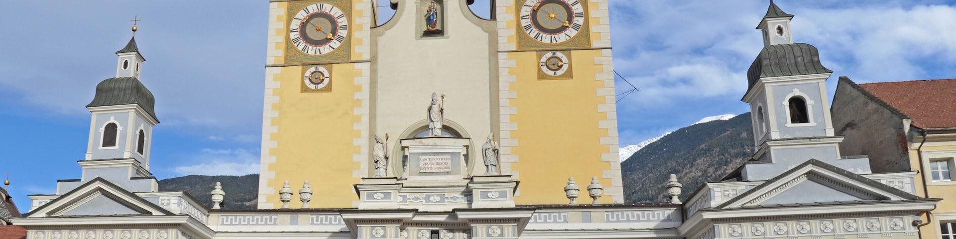 Una iglesia histórica con dos torres y una cúpula central, que presenta un gran reloj y estatuas en la fachada.