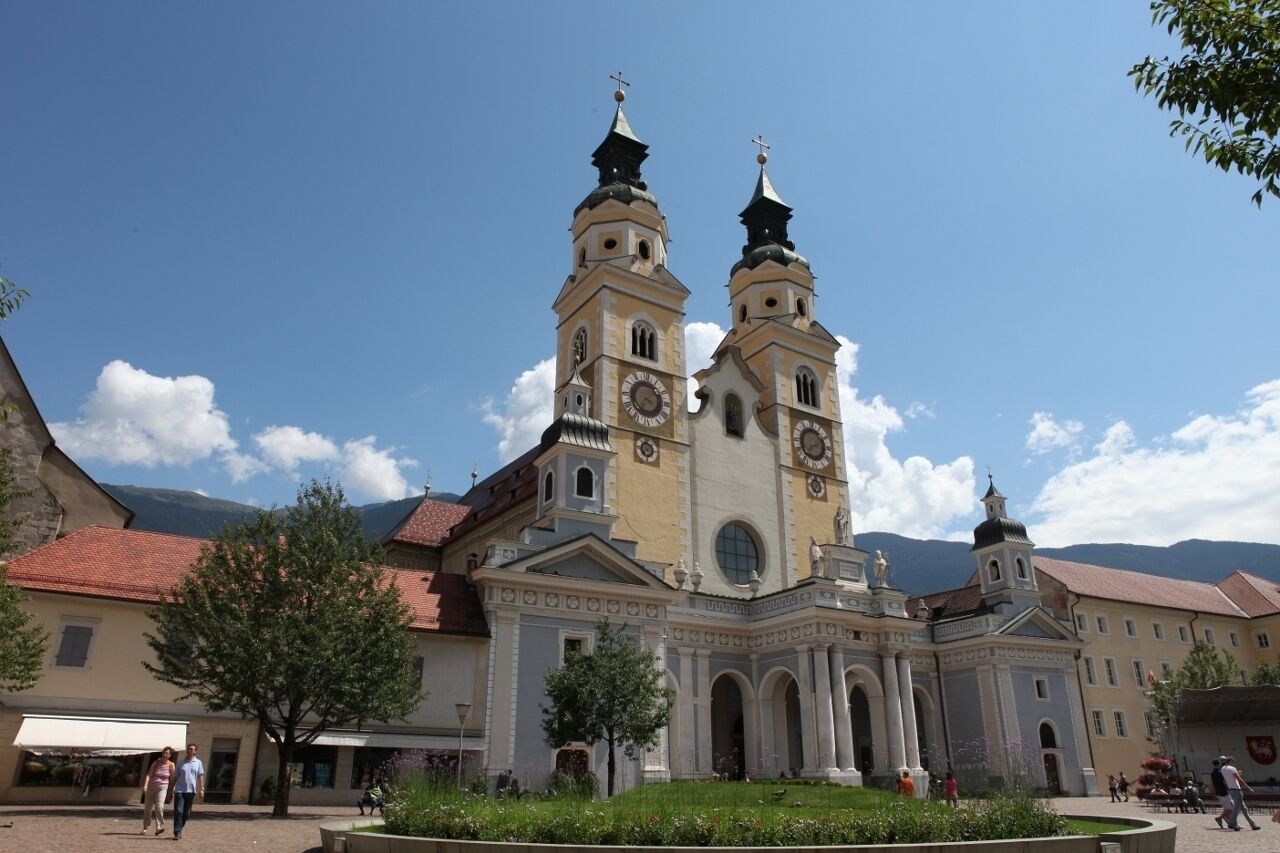 A historic church with two towers and a central dome, surrounded by a town square with buildings and a mountain backdrop.