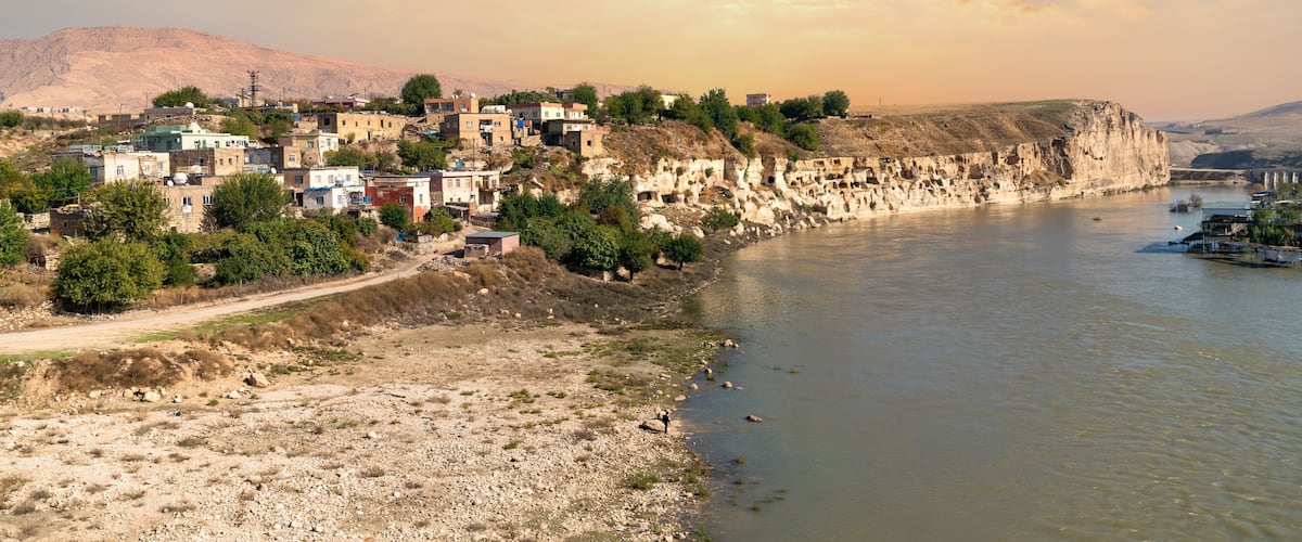A riverside settlement with buildings on a cliff, a river, and mountains in the background.