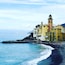 Un pueblo costero con edificios coloridos, una iglesia destacada con un campanario y un litoral rocoso donde la gente disfruta de la playa.