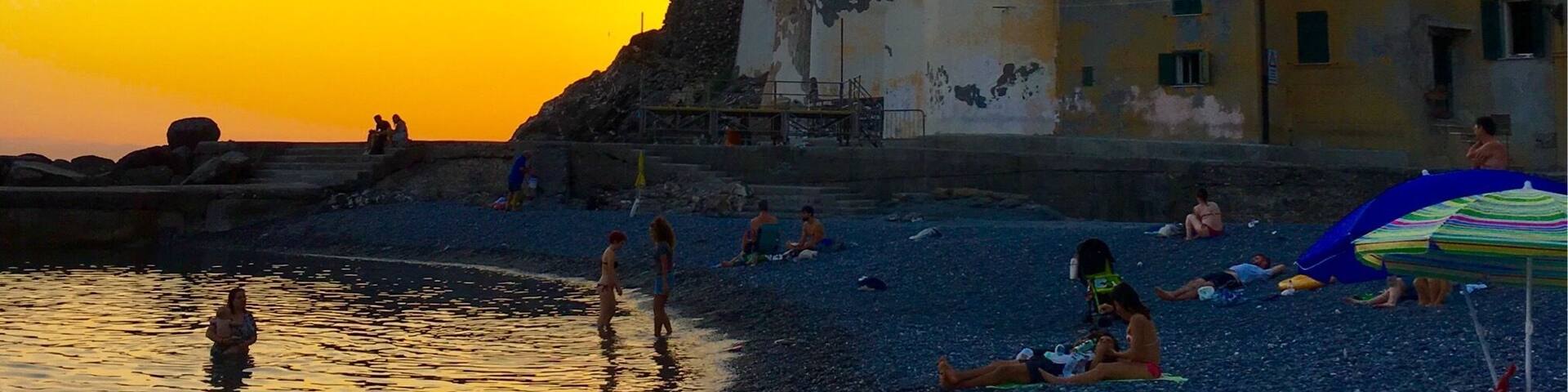 A beach with people at sunset, a church with a bell tower, and buildings in the background.