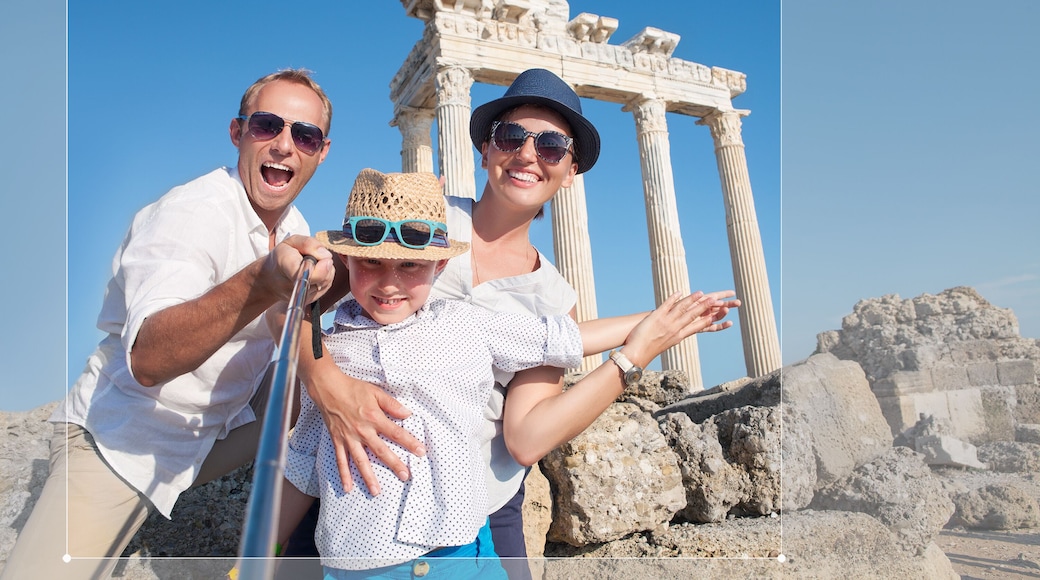 Happy family making a selfie photo using a samrtphone and Selfie stick on summer vacation in Turkey, Side, Manavgat with Temple of Apollo ruines on background. Family traveling concept image.
