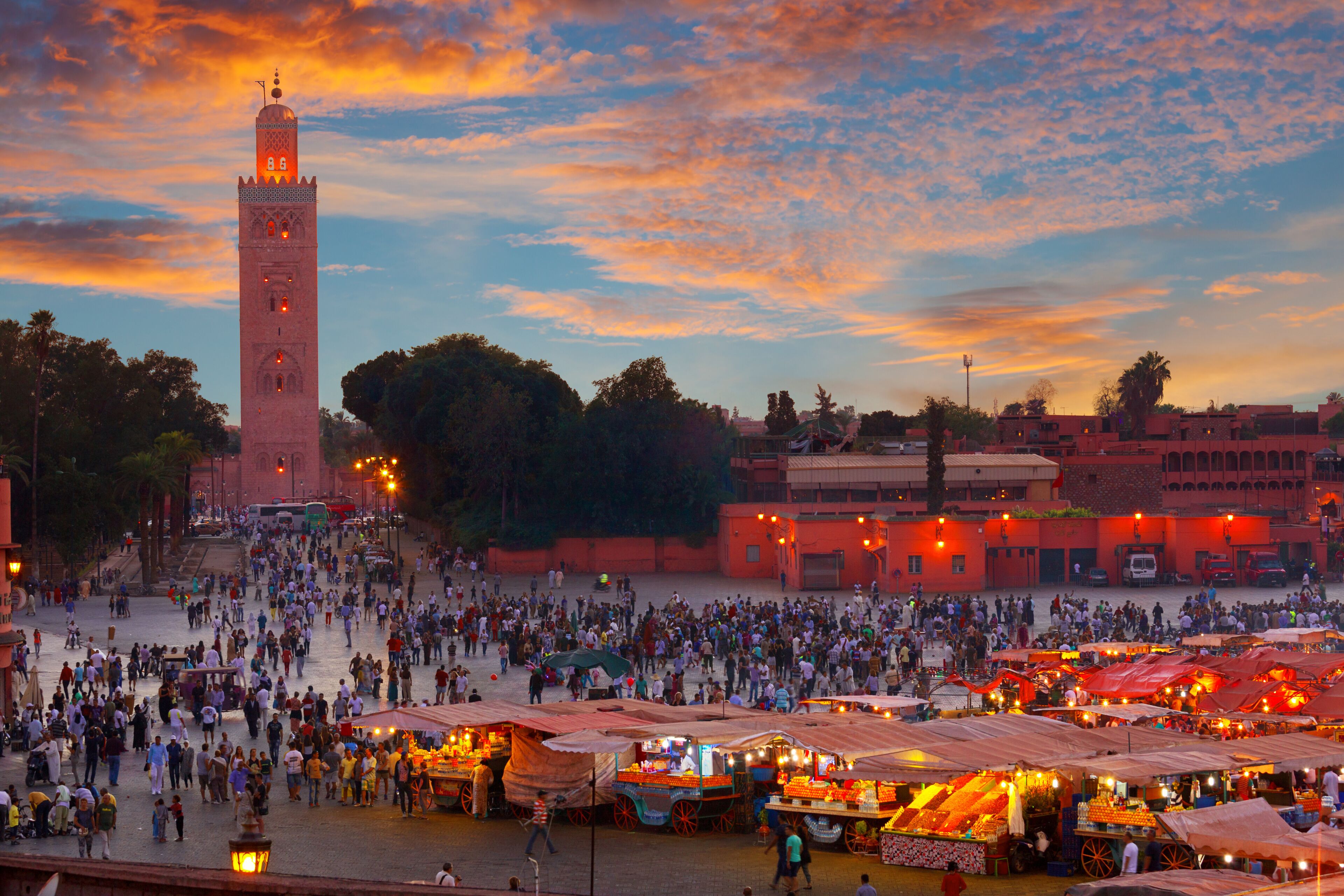 Una plaza animada con una torre destacada, puestos de mercado y un cielo al atardecer.