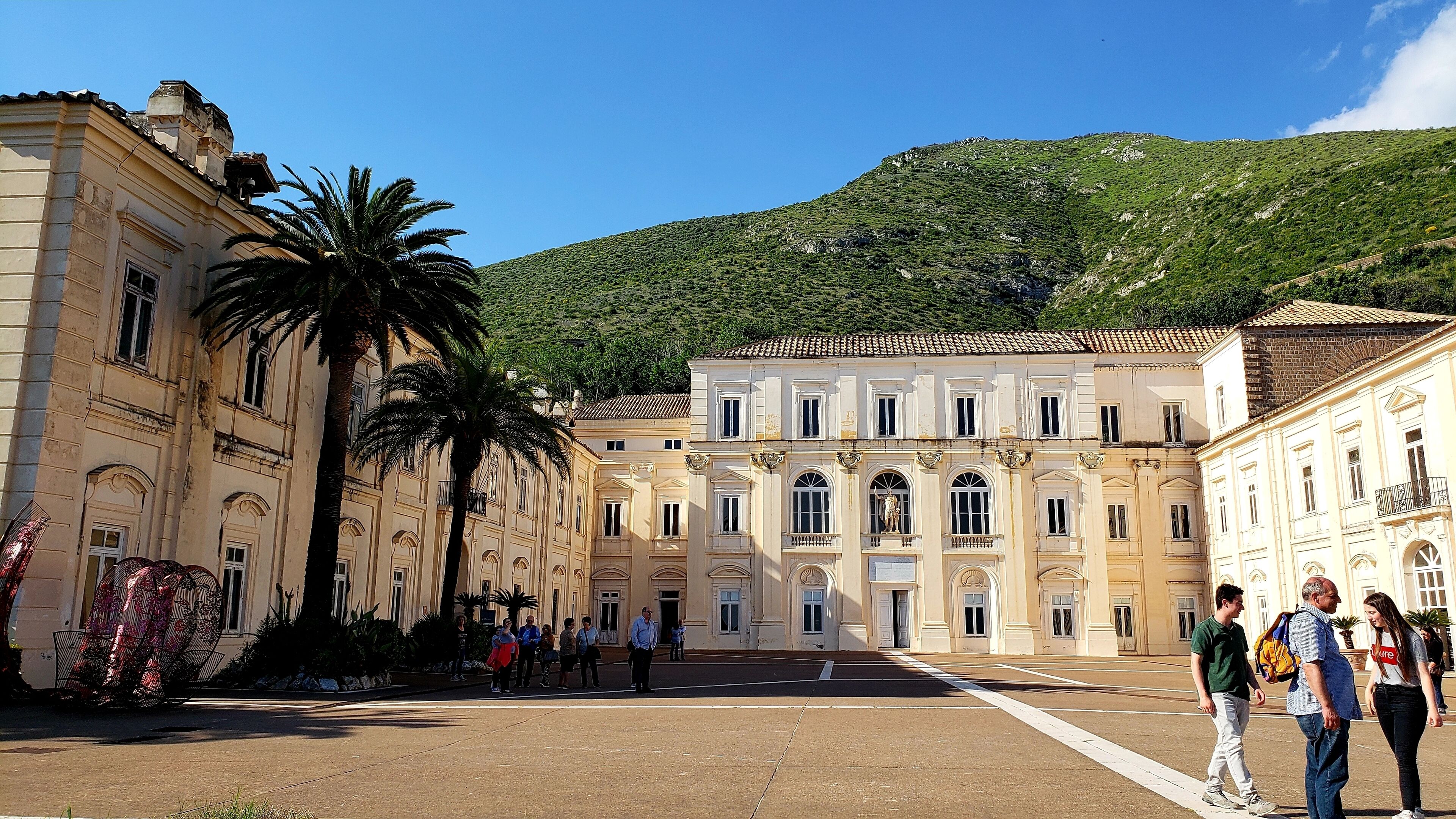 A historic square with classical architecture, palm trees, and a mountainous backdrop.