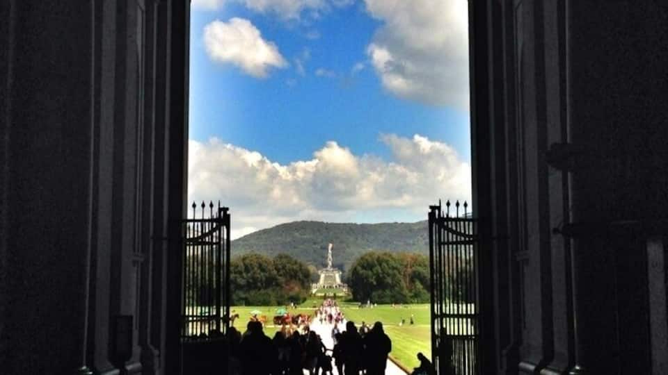 An arched doorway leading to a grassy area with a crowd of people and a statue in the distance.
