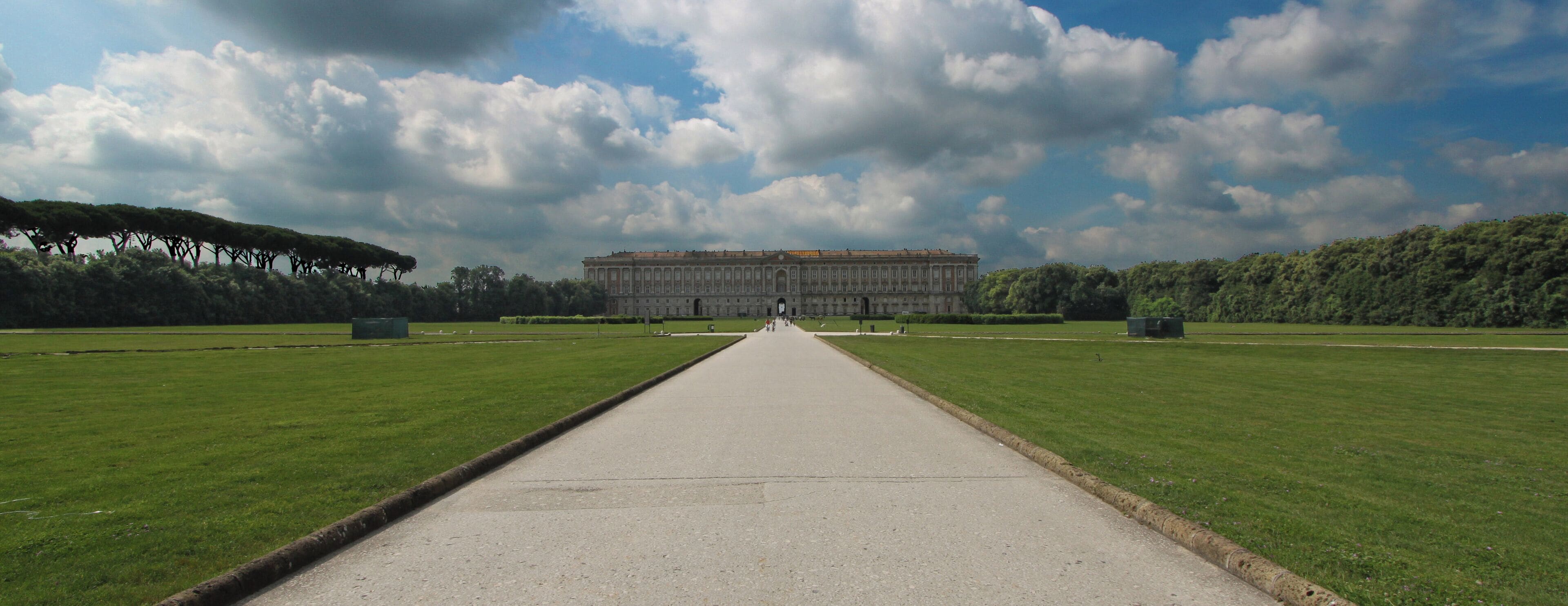 A long, straight pathway leading to a large, classical building with columns, surrounded by well-maintained lawns and trees.