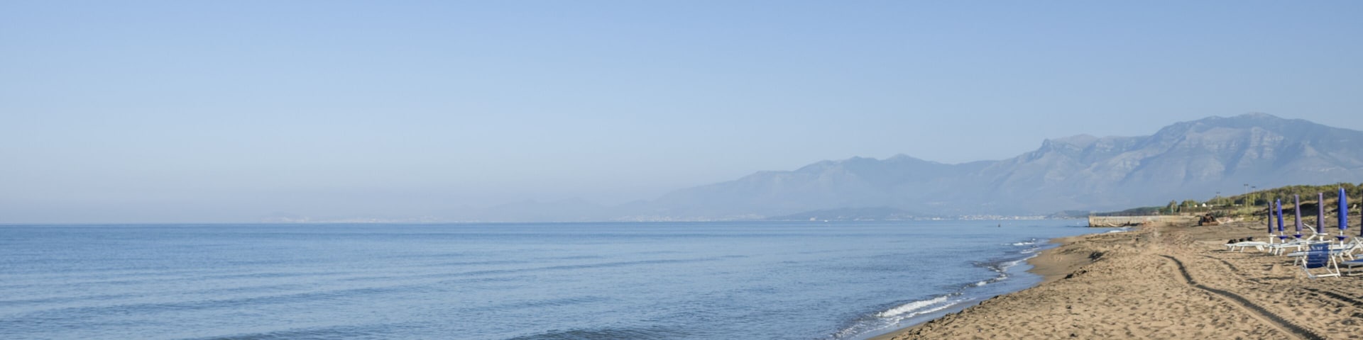 A sandy beach with a calm sea, a clear sky, and distant mountains.