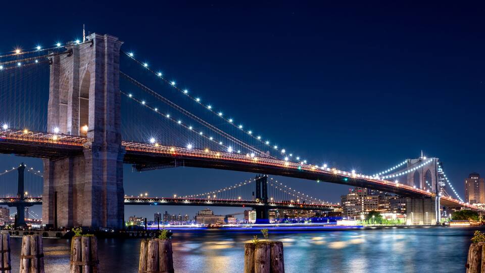 A nighttime view of a large suspension bridge with illuminated cables, a city skyline in the background, and a calm river below.