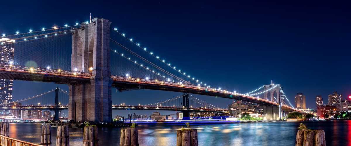 A nighttime view of a large suspension bridge with illuminated cables, a city skyline in the background, and a calm river below.
