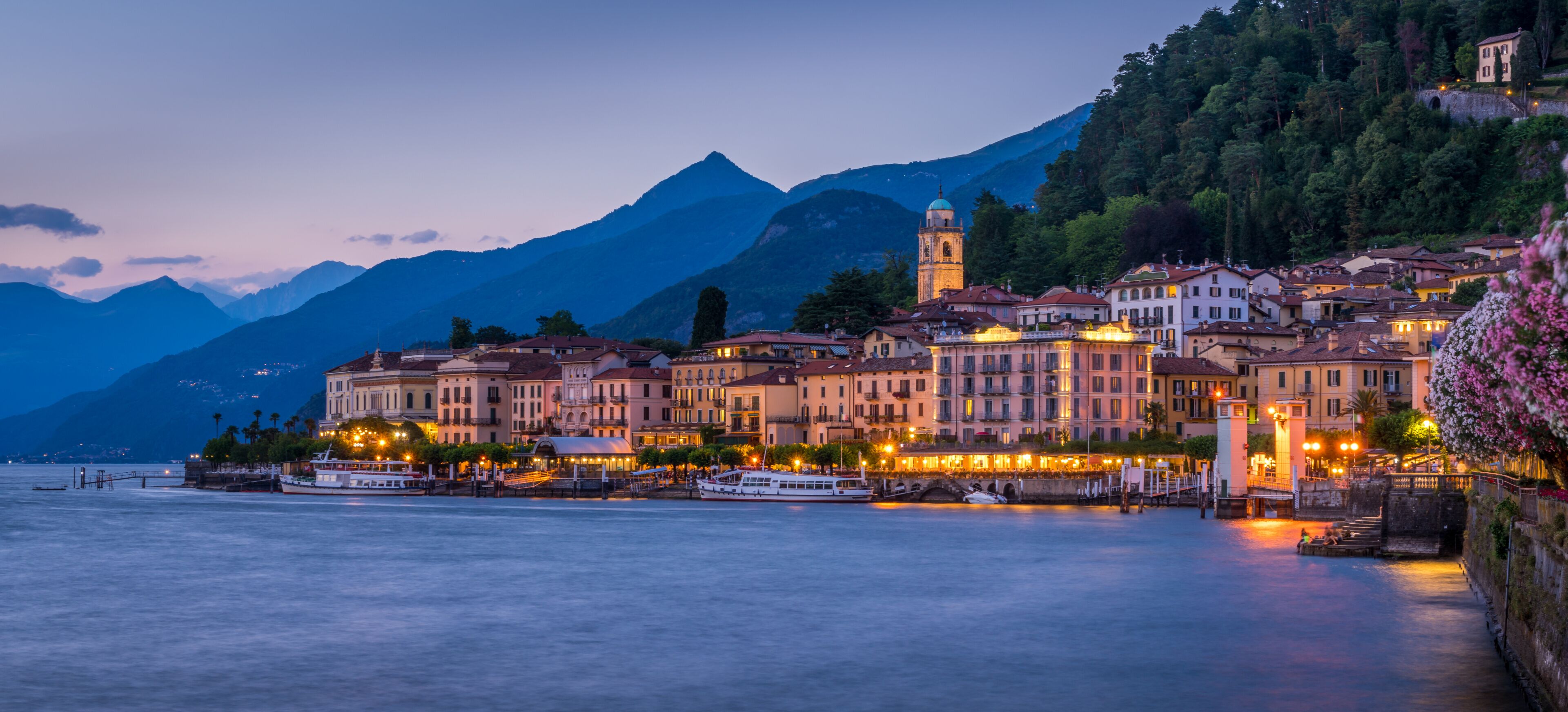 Un pueblo a orillas del lago con edificios históricos, una torre destacada y montañas al fondo.
