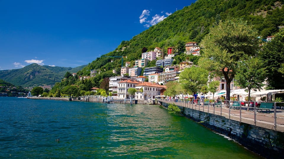 A waterfront town with buildings along a stone embankment, a bridge, and a mountain in the background.