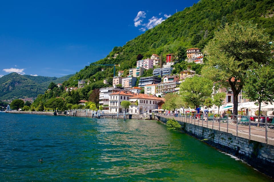 A waterfront town with buildings along a stone embankment, a bridge, and a mountain in the background.