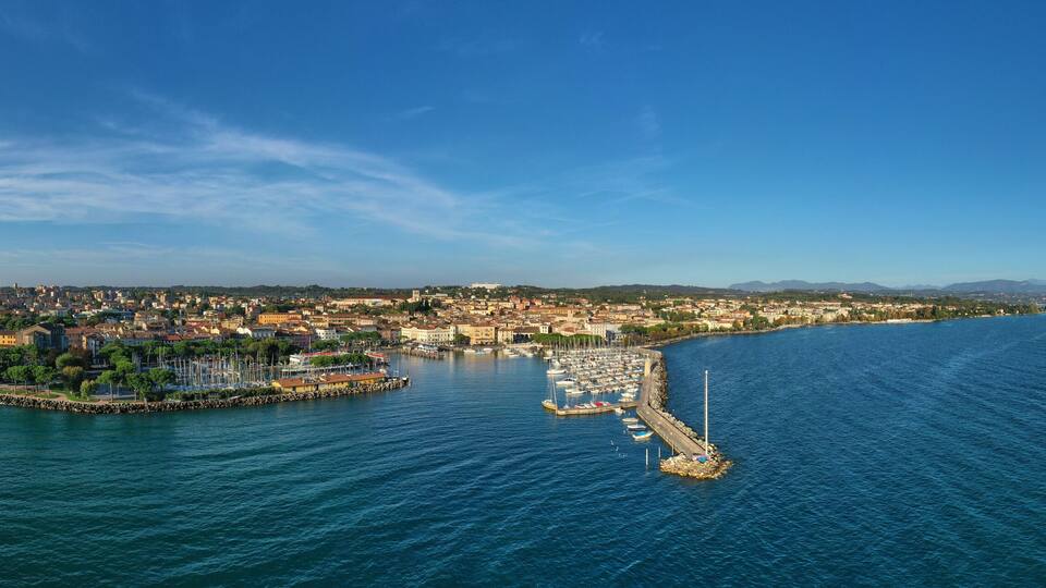 A coastal cityscape with a marina, a rocky breakwater, and a distant mountain range.