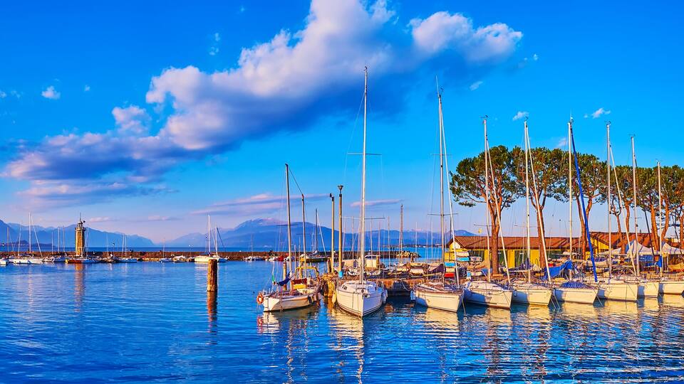 A marina with boats, a lighthouse, and mountains in the background.