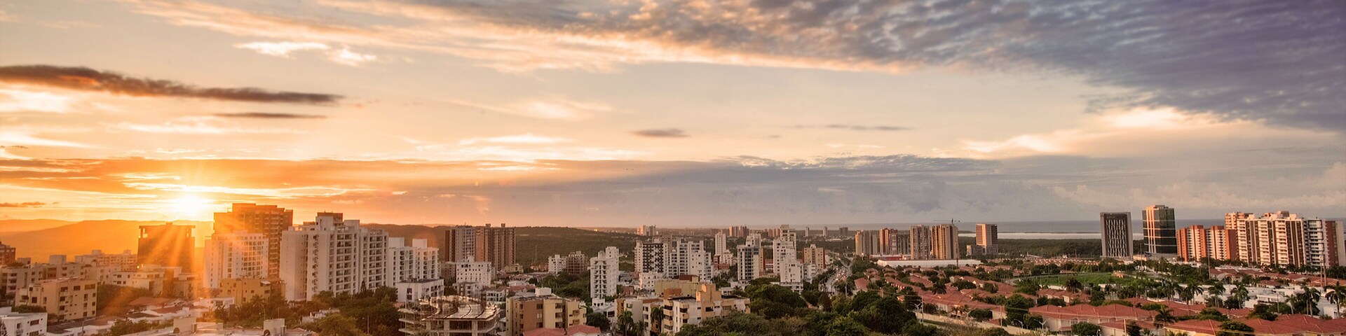 A cityscape at sunset with a mix of residential and high-rise buildings, a clear sky, and the sun setting on the horizon.