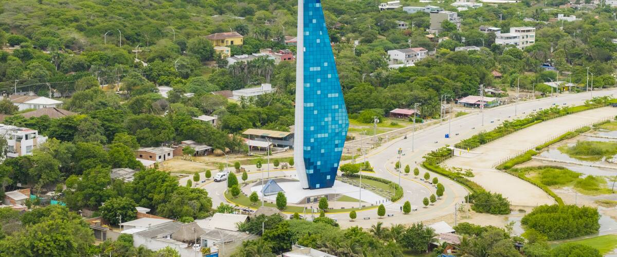 A modern skyscraper with a blue facade, surrounded by lush greenery and a small town with winding roads.