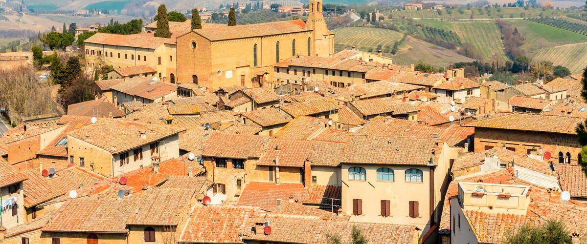 A town with terracotta-roofed buildings, a prominent church with a bell tower, and rolling hills in the background.