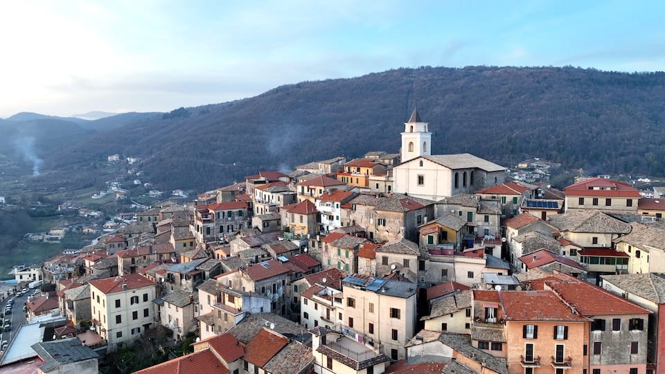 A densely packed hillside town with historic buildings, a prominent church tower, and a mountainous backdrop.
