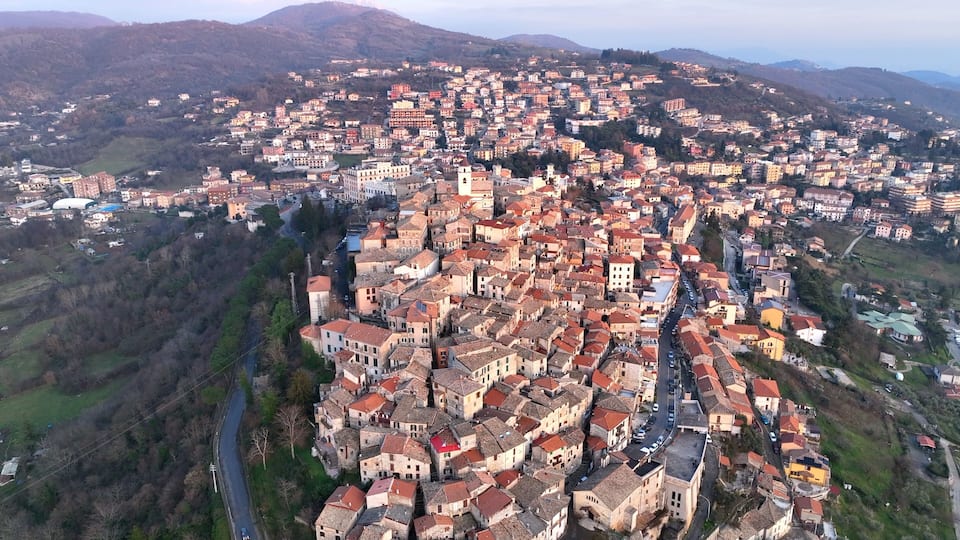 A densely packed town with red-roofed buildings, surrounded by hills and a river.
