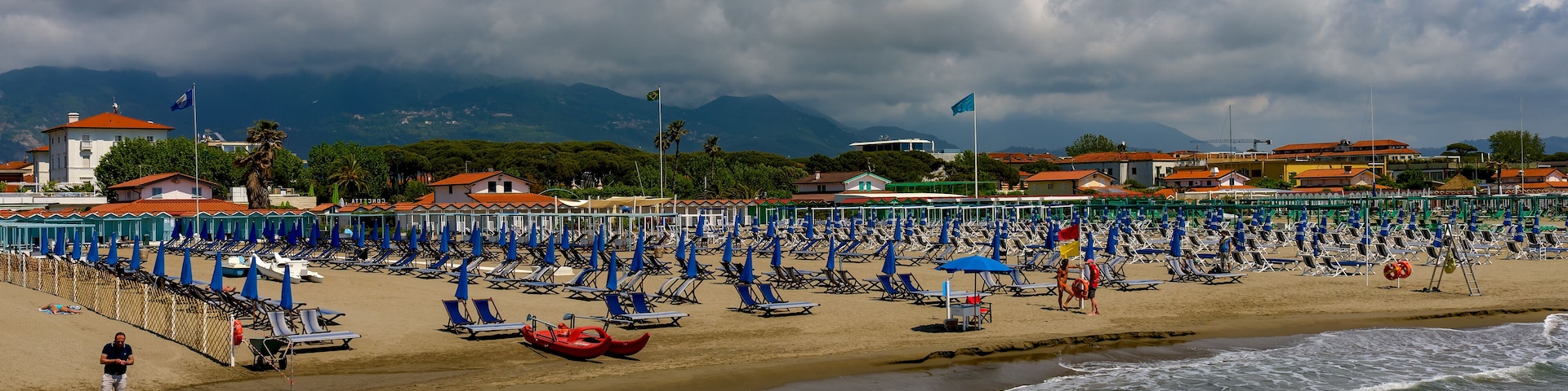 A beach with rows of blue umbrellas and lounge chairs, a rocky shoreline, and a mountainous backdrop.