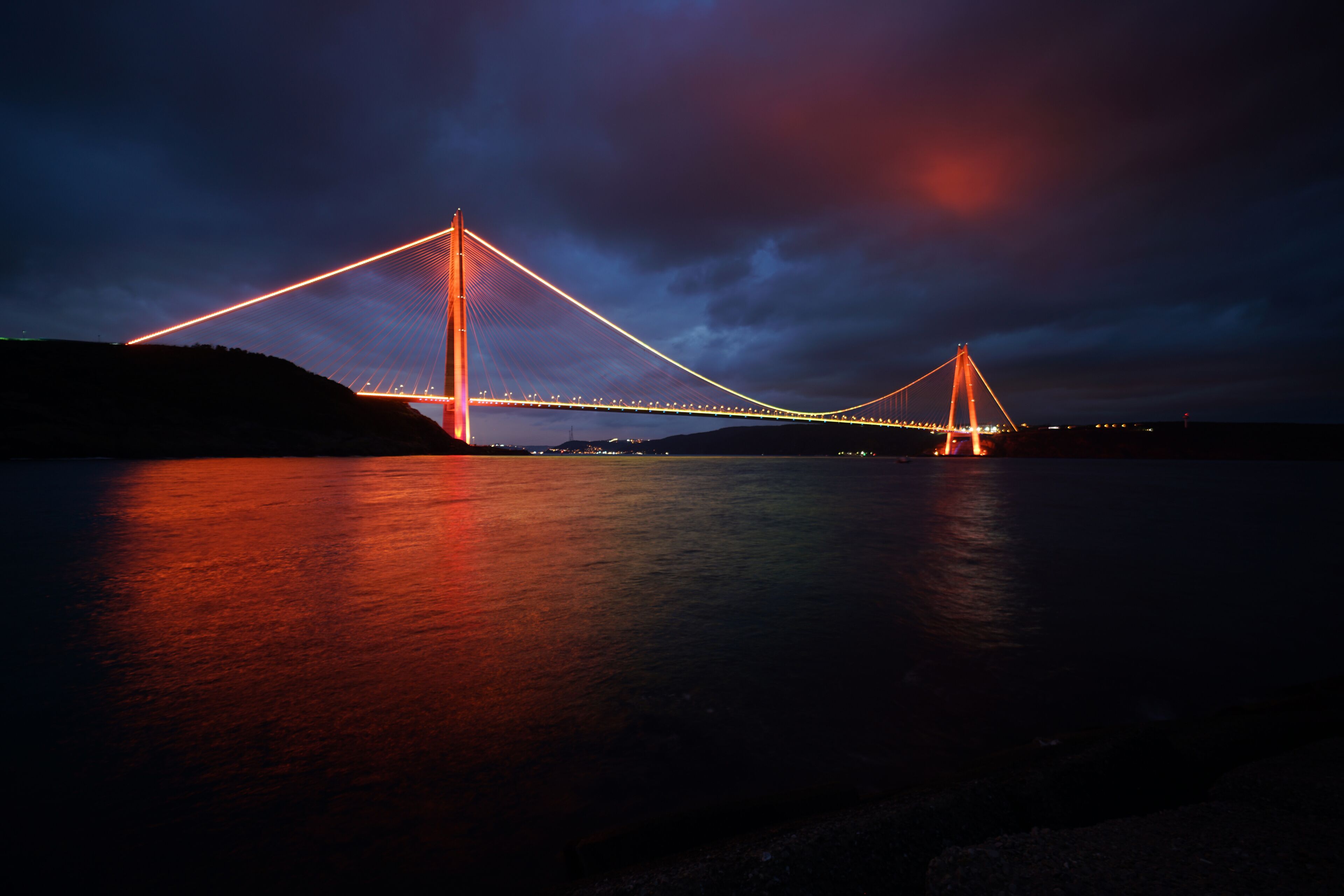                Osmangazi Bridge. The photo of the bridge taken from the front while its colorful lights are on at night. The bridge lights are reflected on the sea, creating a magnificent view.