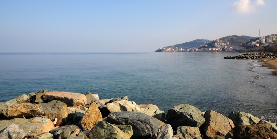 A coastal landscape with large rocks in the foreground, calm water, and a city on a hill in the background.