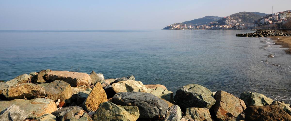 A coastal landscape with large rocks in the foreground, calm water, and a city on a hill in the background.