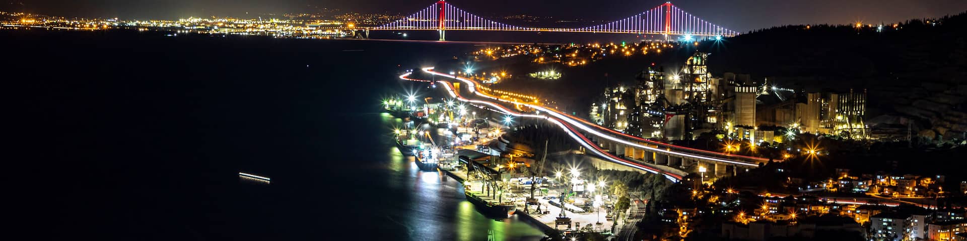 A coastal city at night with a brightly lit bridge, a winding road along the coastline, and a harbor area with boats.