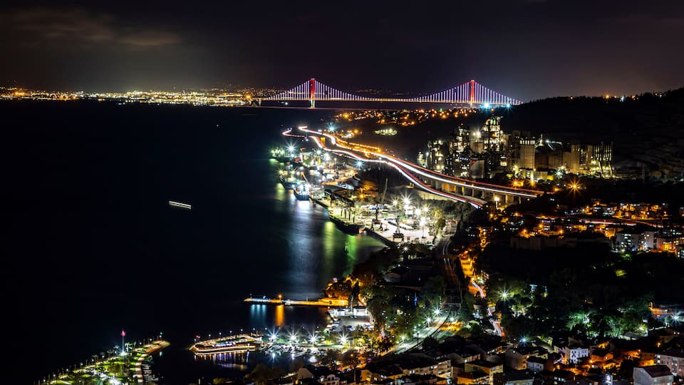 Uma cidade costeira à noite, com uma ponte bem iluminada, uma estrada sinuosa ao longo da costa e uma área portuária com barcos.