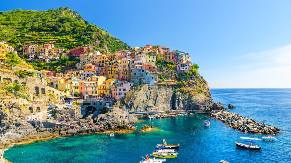Manarola traditional typical Italian village in National park Cinque Terre, colorful multicolored buildings houses on rock cliff, fishing boats on water, blue sky background, La Spezia, Liguria, Italy
