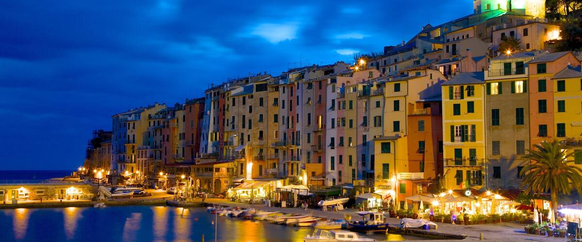 A waterfront cityscape at night with colorful buildings, a prominent church tower, and boats docked along the quay.