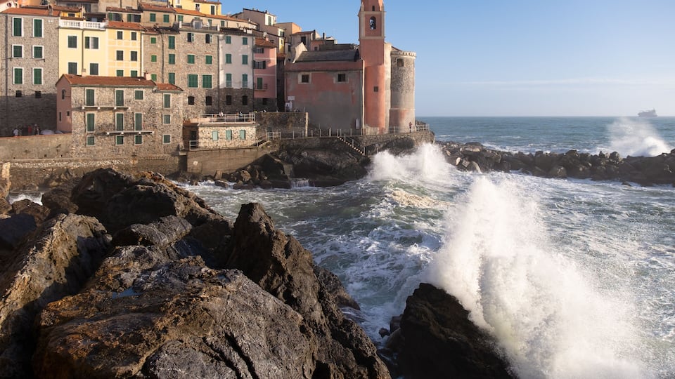 A coastal town with historic buildings, a prominent lighthouse, and rough sea waves crashing against the rocks.