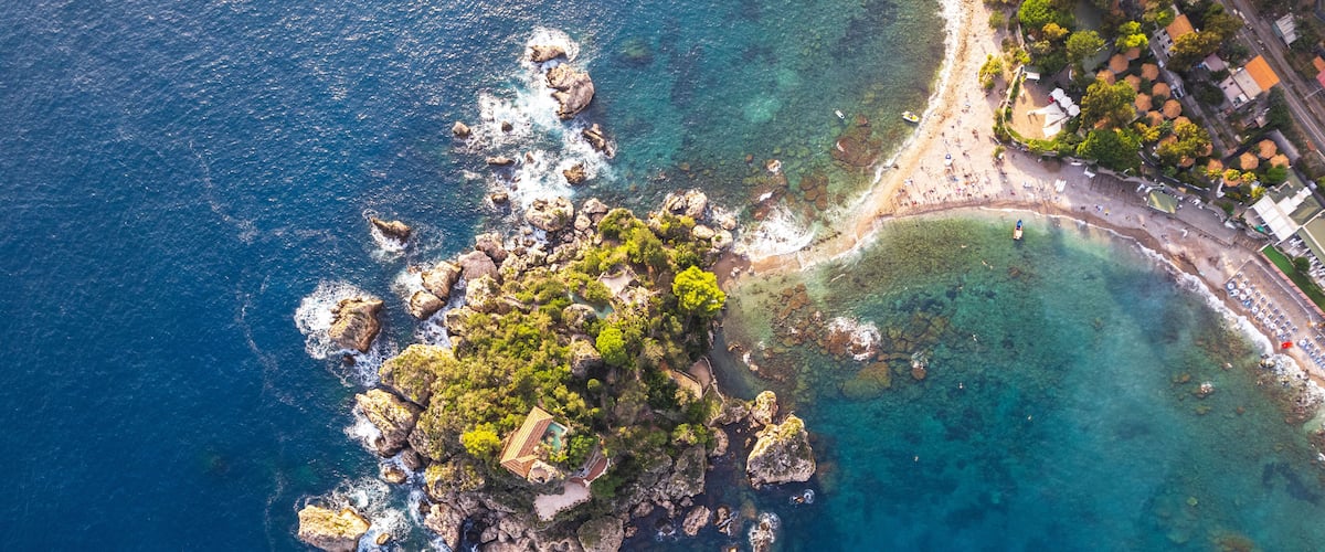 An aerial view of a rocky island with clear blue waters, surrounded by a coastal town with buildings and a beach.