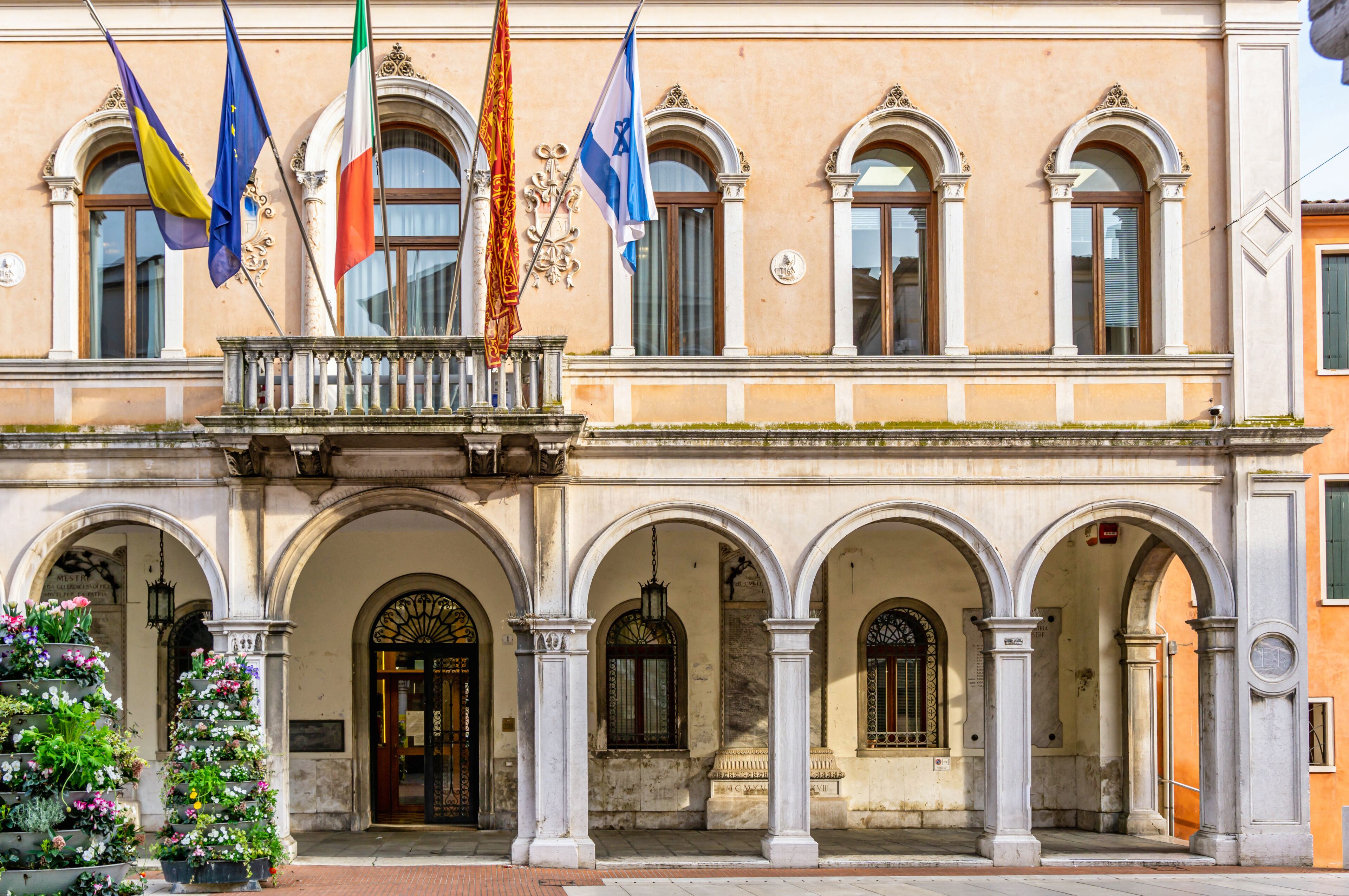 A historic building with multiple arches and flags, including the European Union and national flags.