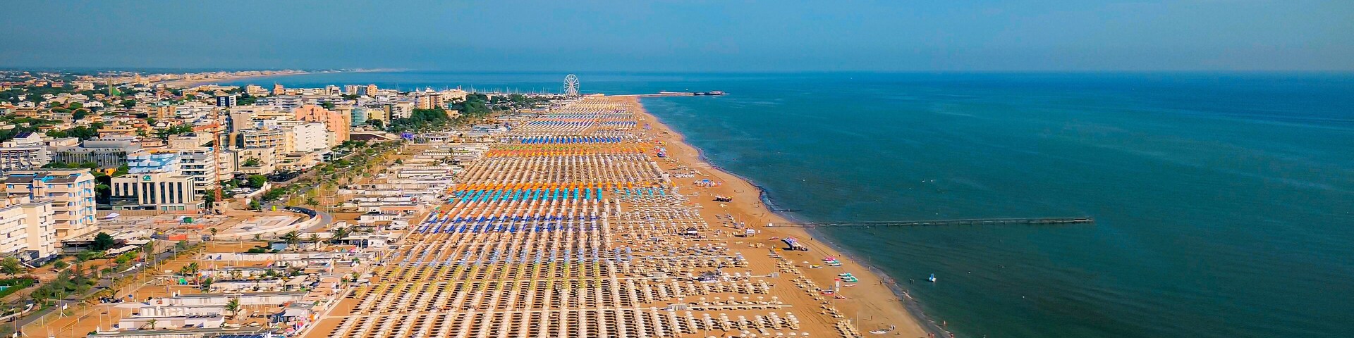 Un paisaje urbano costero con una playa larga bordeada por filas de tumbonas y sombrillas, un muelle que se adentra en el mar, y una noria a lo lejos.