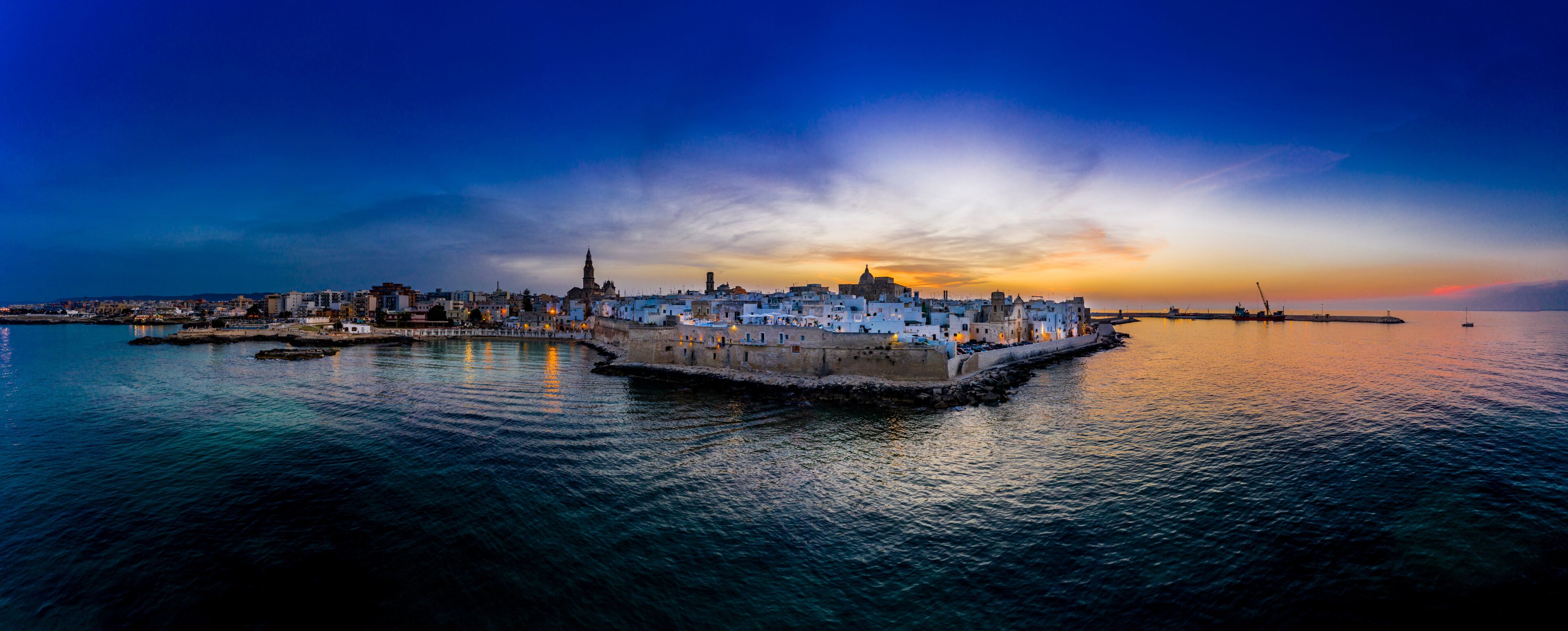 A coastal cityscape at sunset with a prominent tower and a calm sea.