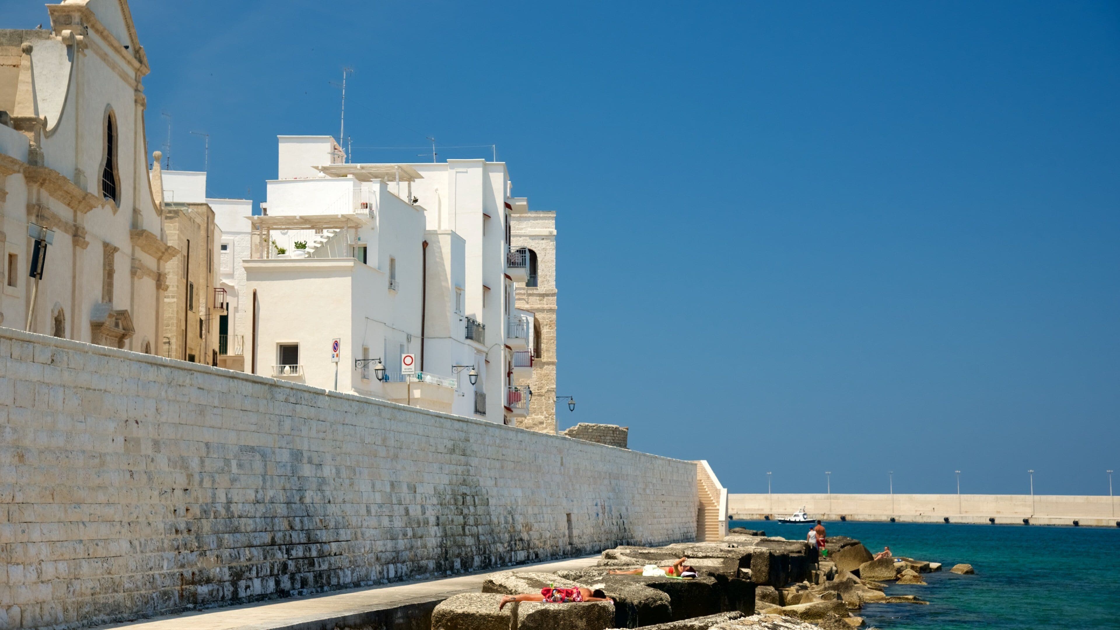 A coastal cityscape with white buildings, a stone wall, and a clear blue sky.