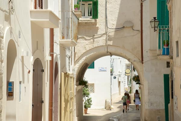 Uma rua estreita com um arco, edifícios com persianas e uma placa com 'Cacace'.
