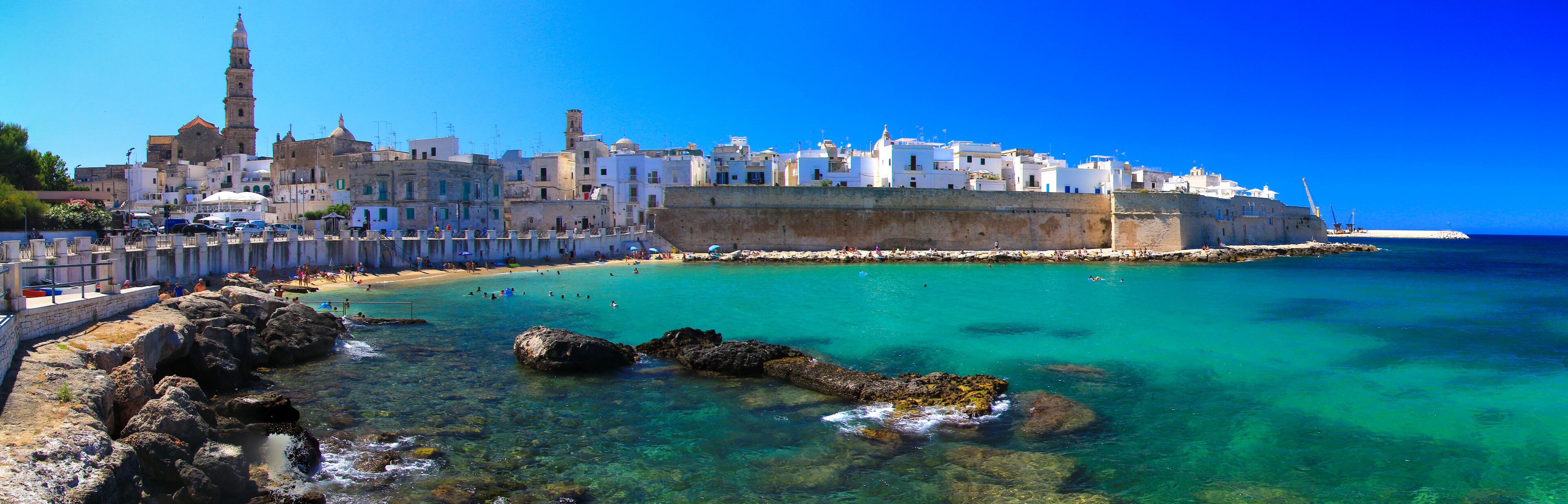 A coastal town with clear turquoise waters, a sandy beach, and historic buildings including a prominent tower.